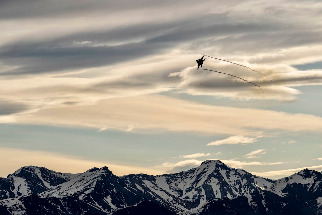An Air Force jet flies above a snow-capped mountain range in blue- and pink-streaked sky.