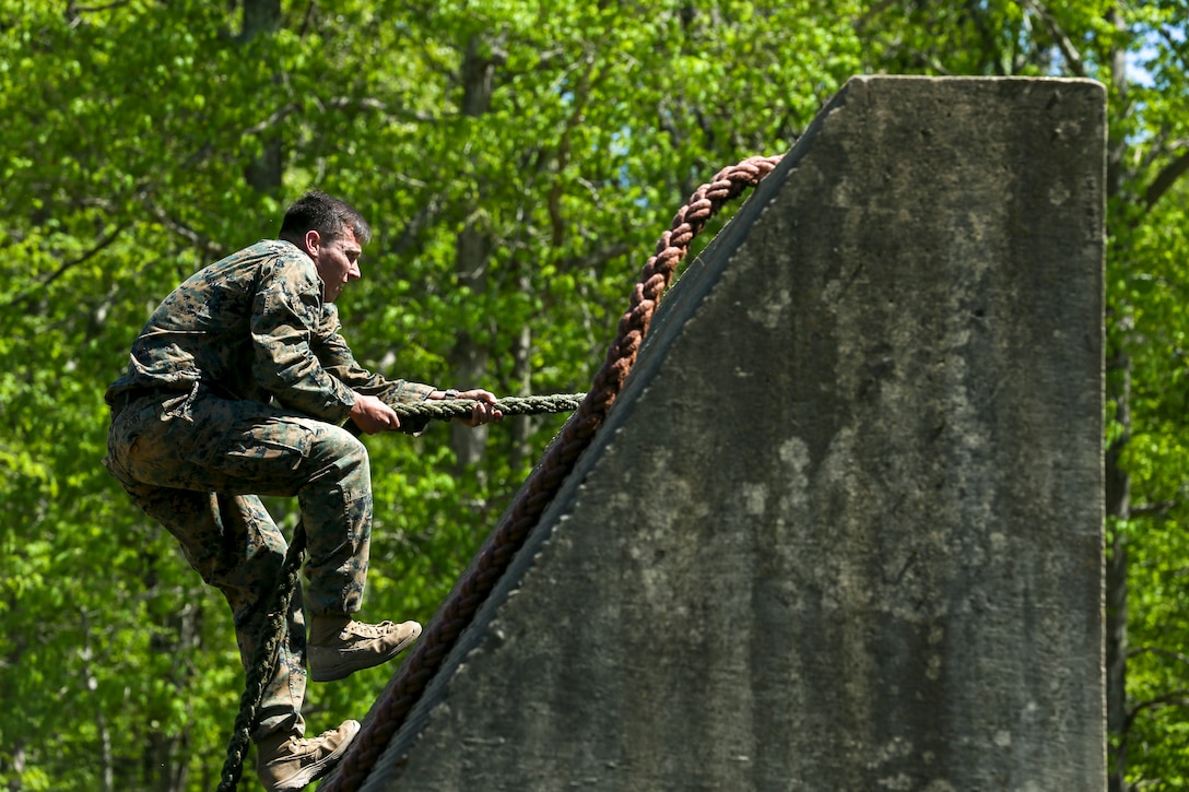 A Marine uses a rope to scale a slanted concrete-type wall with trees in the background.