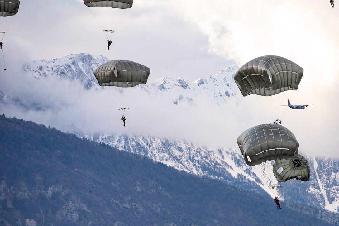 Soldiers parachute against a backdrop of snowy mountains, with an aircraft flying in the distance.