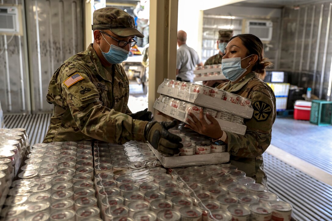 Two soldiers handle cases of canned food in a large warehouse-type room.