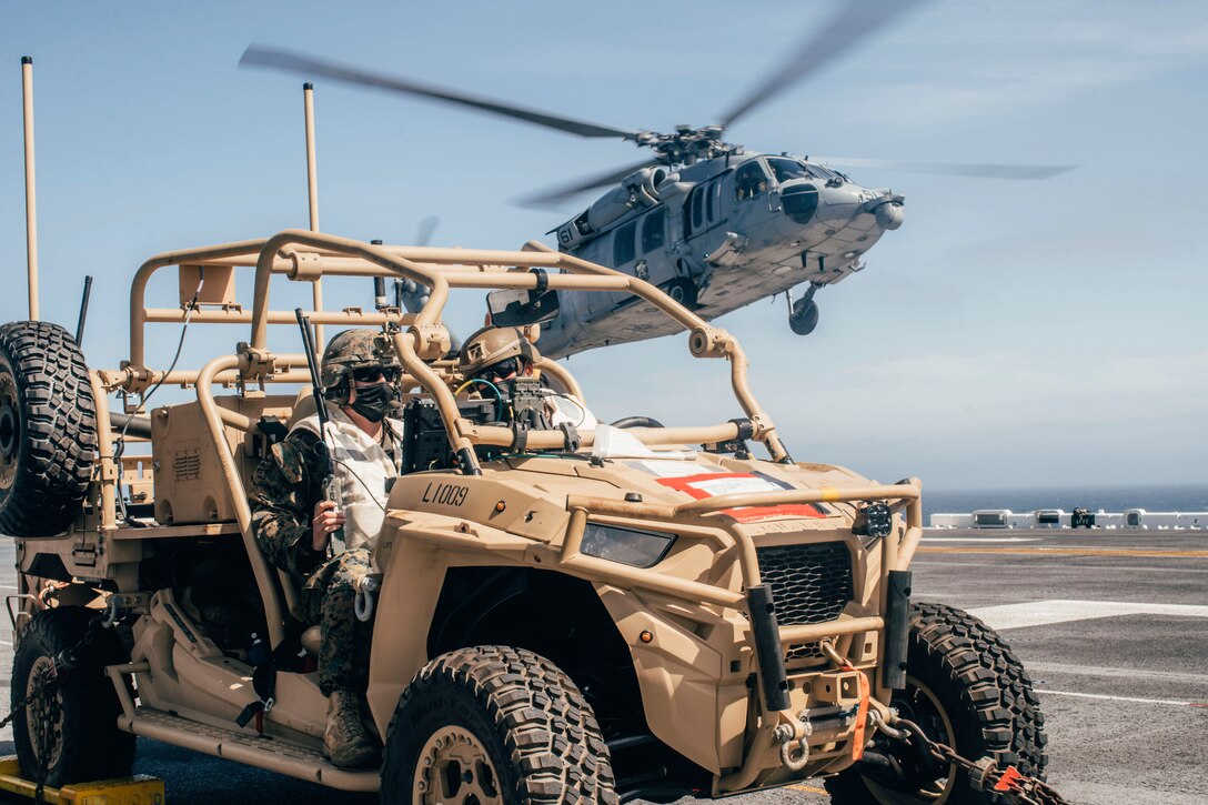 Marines sit in a military vehicle while a helicopter takes off from a ship.