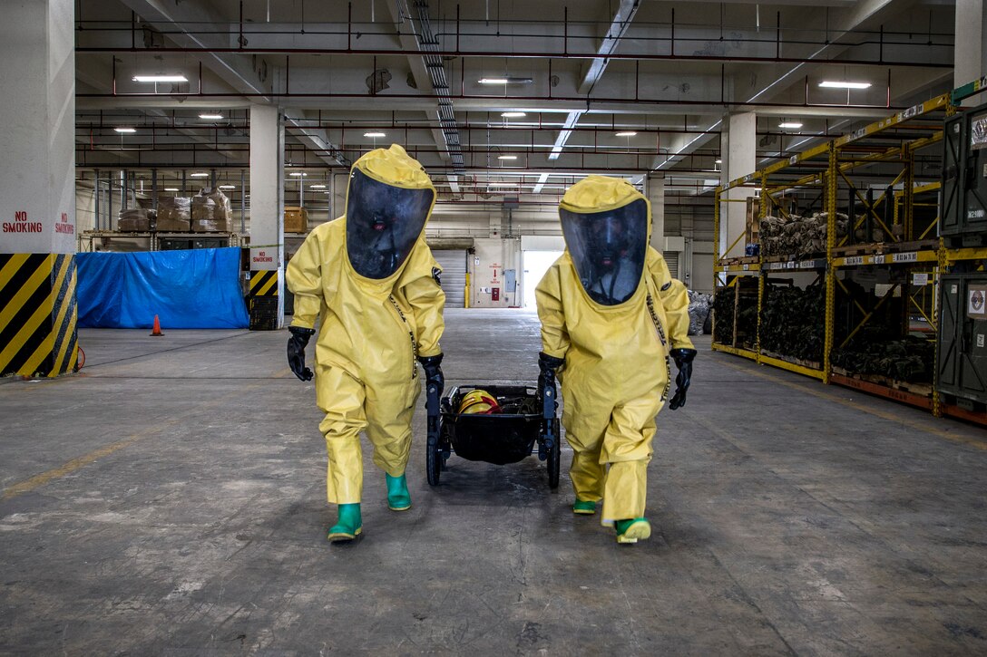 Two Marines wearing full-body yellow protective gear walk through a warehouse-type facility.