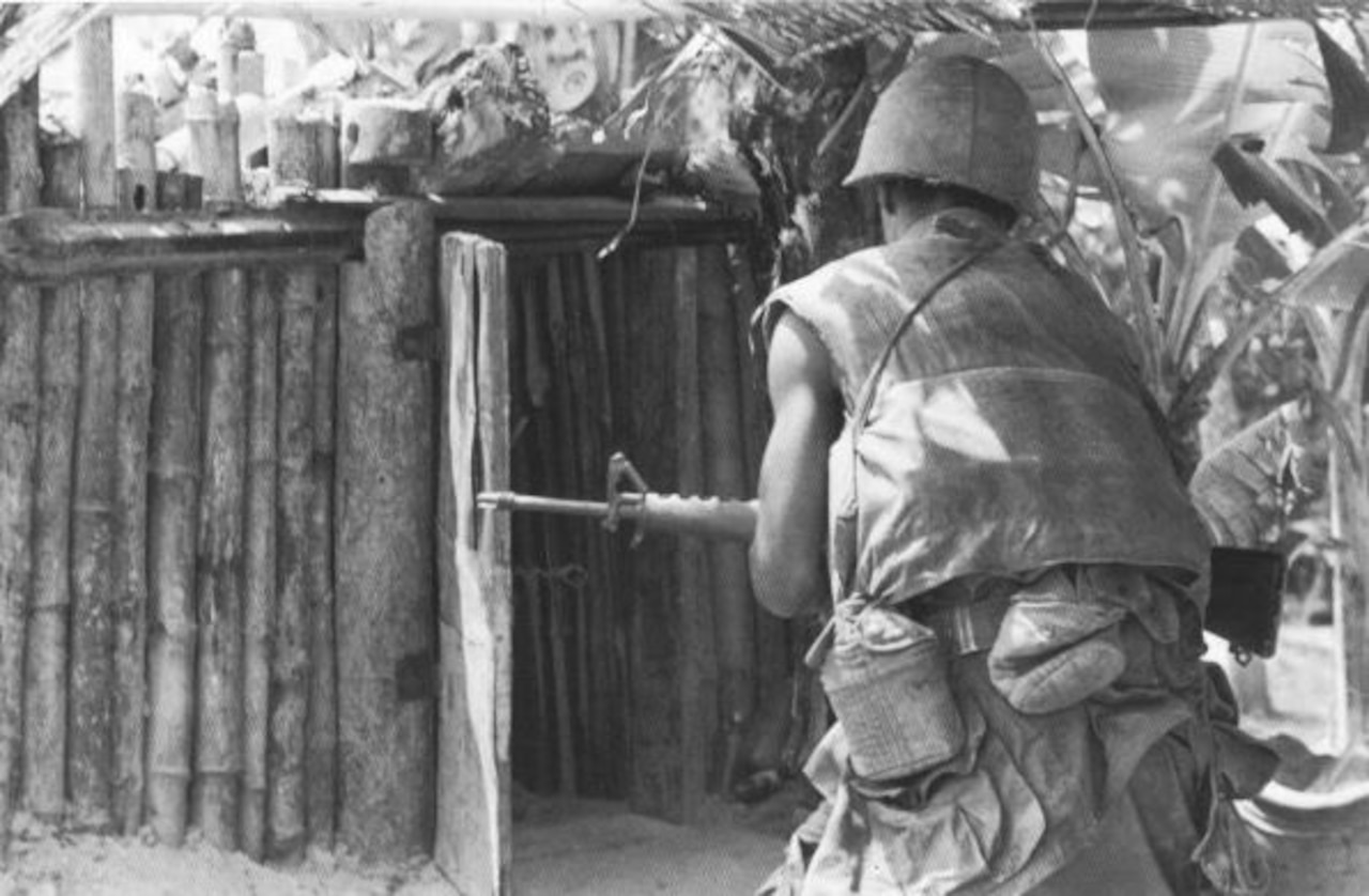 A man in a combat uniform and carrying a rifle approaches the entrance to a hut.