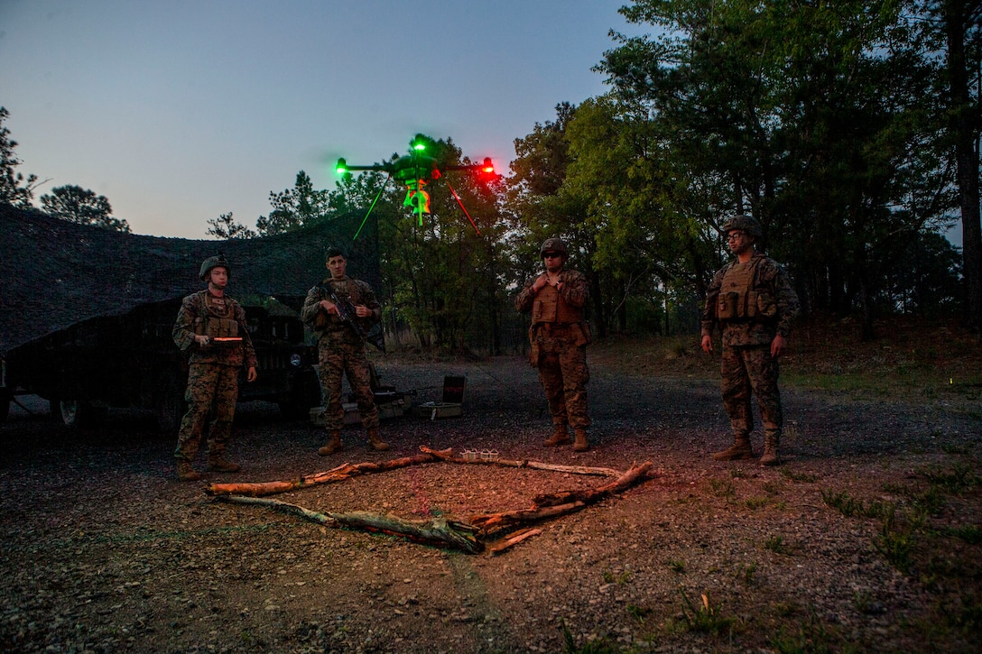 Four Marines stand and watch as a small lighted drone hovers over a square patch of ground demarcated by sticks against a dark sky.
