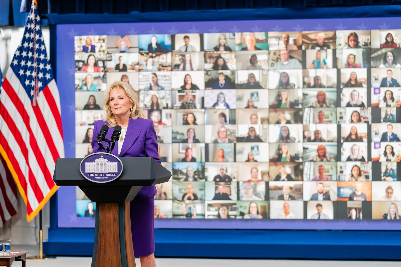 A woman in a suit stands behind a lectern with microphones. A sign on the lectern indicates that she is at the White House. A collage of monitors is behind her.