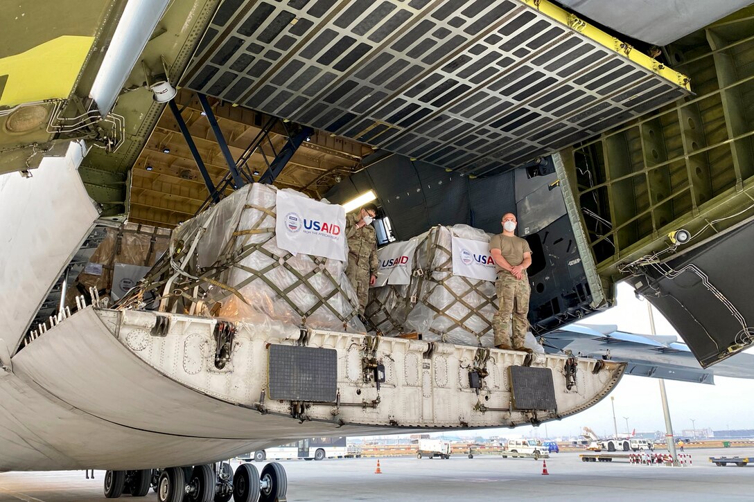 Service members stand by pallets of supplies in the back of an aircraft.
