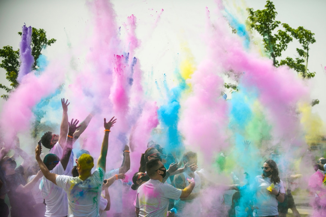 Service members and civilians throw pink, blue and yellow powder into the air while standing outside in a group.