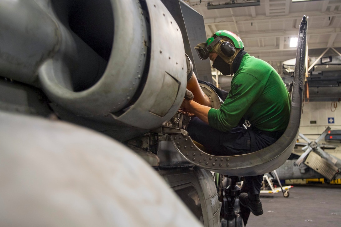 A sailor in a green shirt sits in a curved metal protuberance extending out from an aircraft.