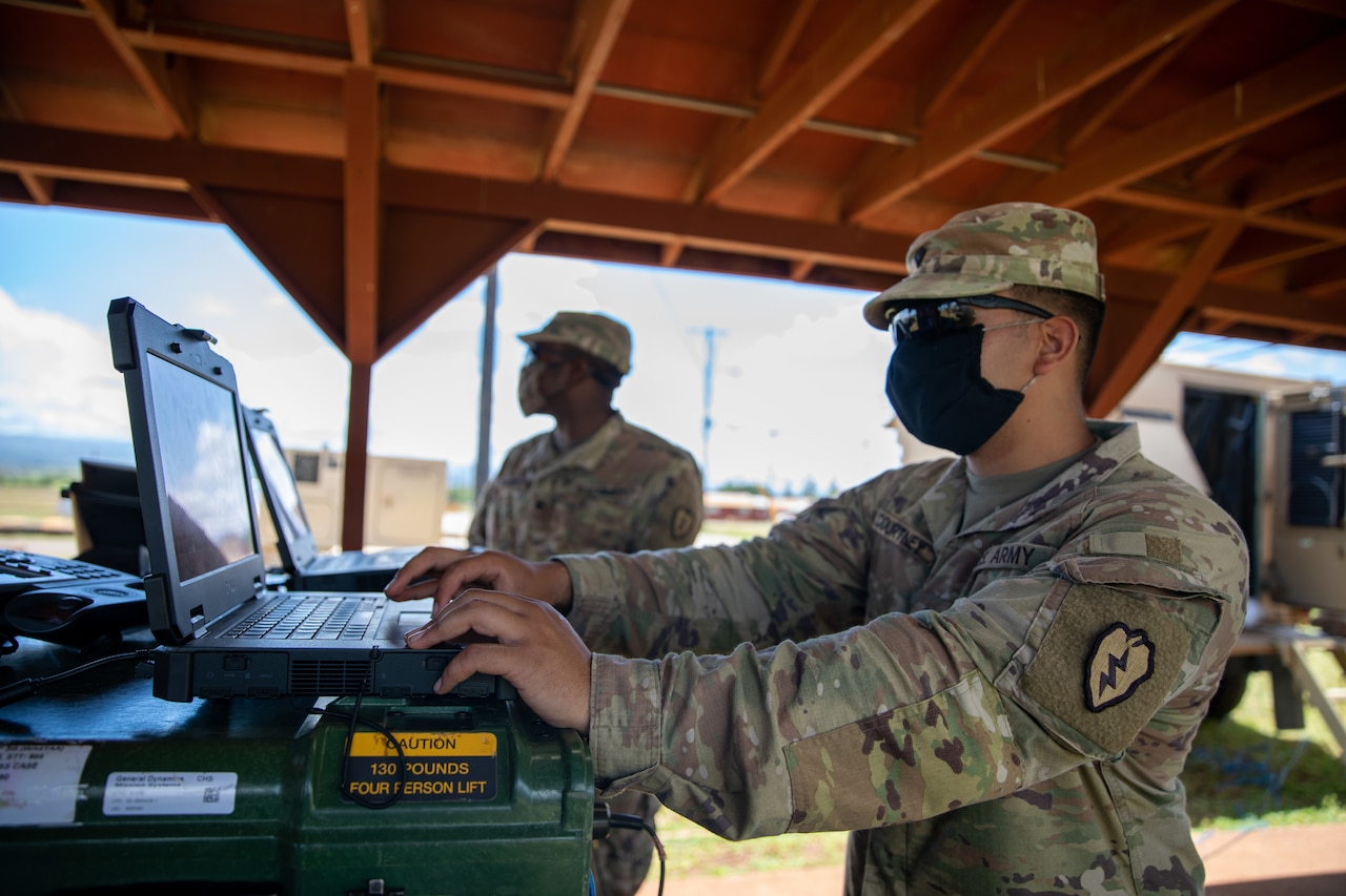 Soldiers work on laptops under a tent outdoors.