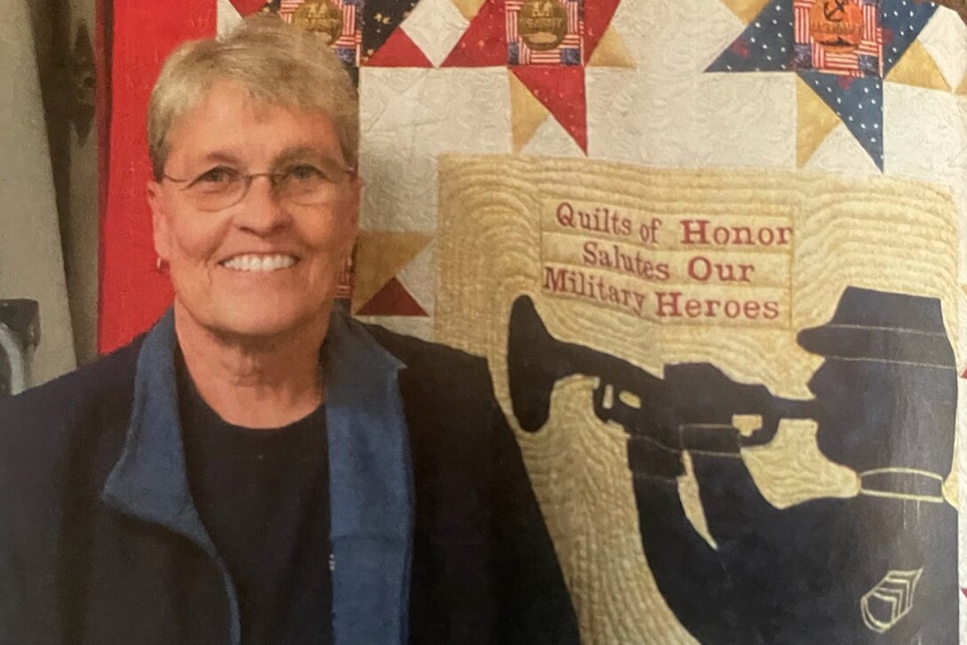 A woman poses for a photo next to a quilt.