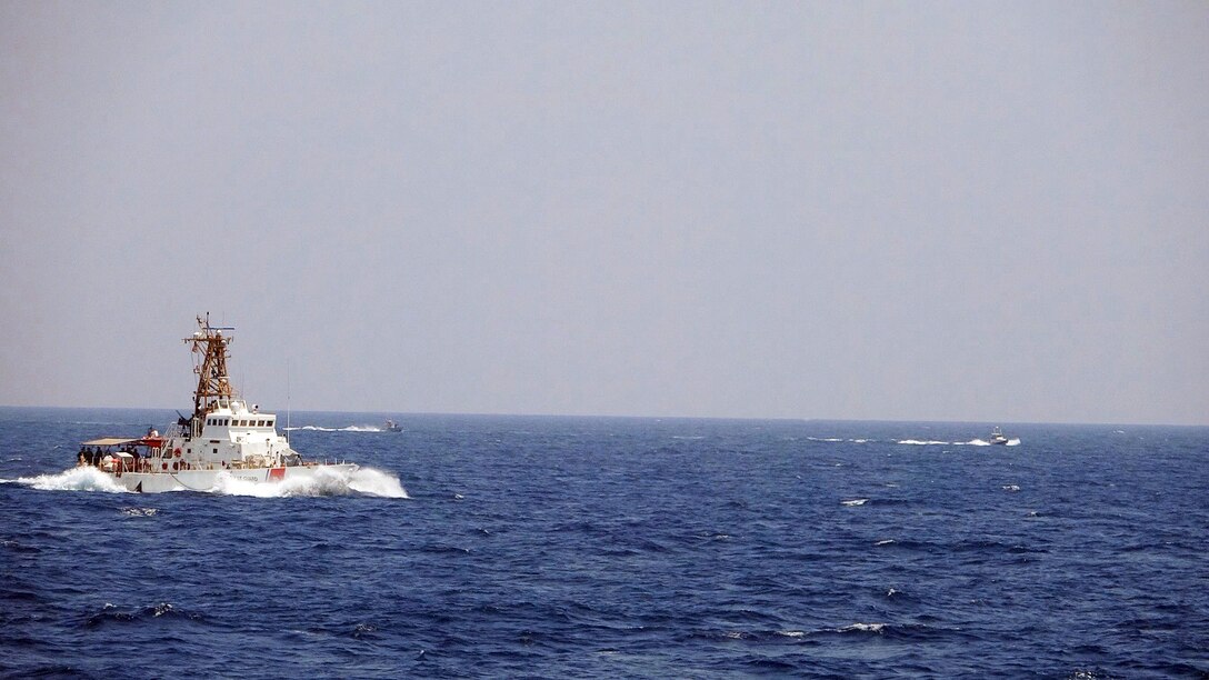A Coast Guard vessel moves through blue ocean waters. In the rear, two smaller speed boats maneuver in the water.