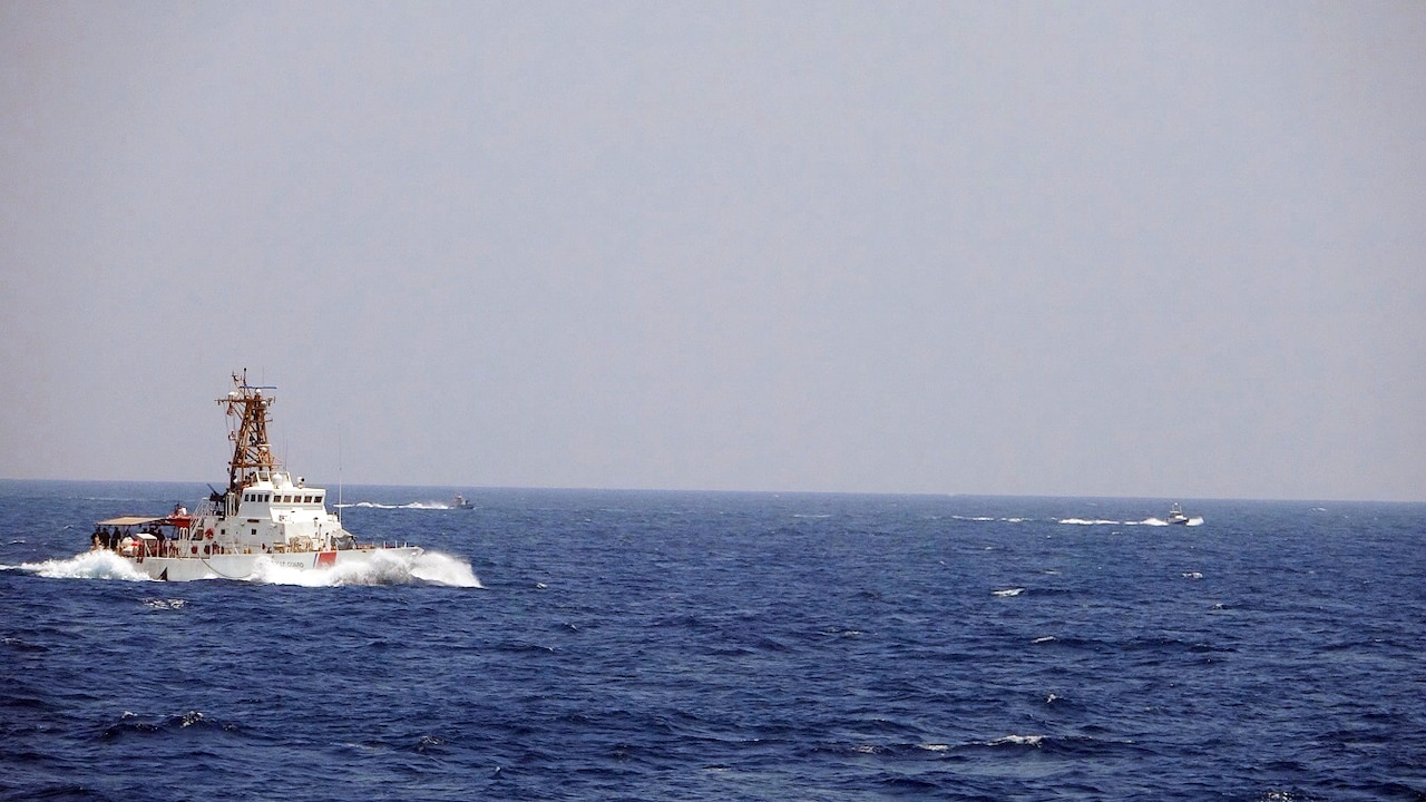 A Coast Guard vessel moves through blue ocean waters. In the rear, two smaller speed boats maneuver in the water.