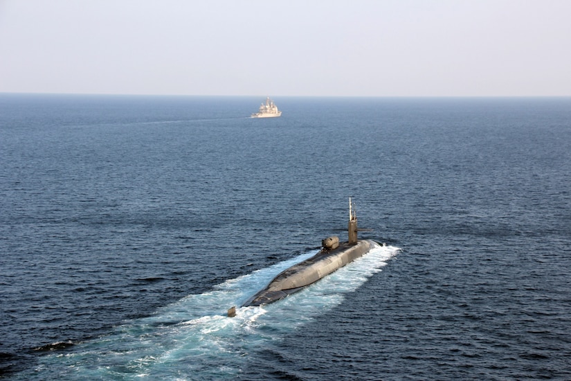 A submarine moves through the water. Far in front of it is a surface vessel.