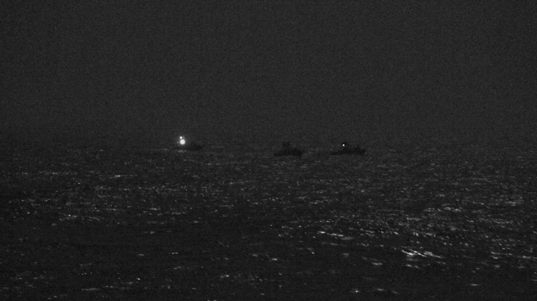 At night, three small boats are sitting near each other. One has a light on.