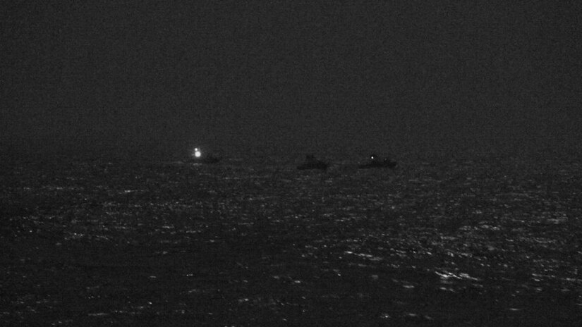 At night, three small boats are sitting near each other. One has a light on.