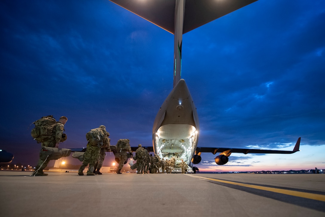 A line of soldiers board a large military aircraft under a dark sky.