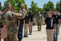 man in uniform with his right hand raised, in front of multiple young people holding up their right hand.