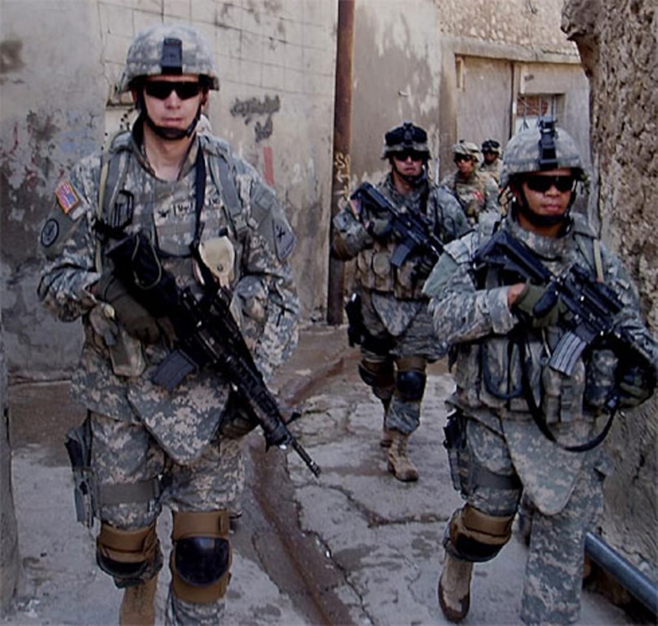 Five soldiers, dressed in combat gear and carrying weapons, walk down a narrow path with buildings on either side.