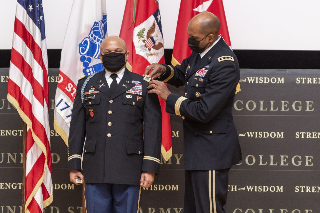 A soldier stands at attention as another soldier pins a rank insignia onto the shoulder of his uniform.