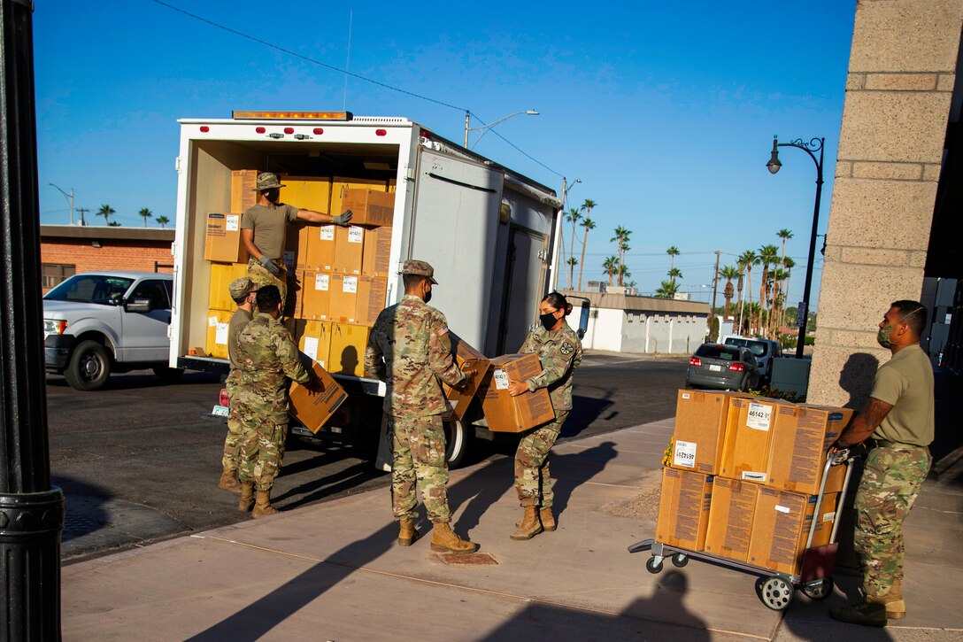 A National Guardsman wearing a face mask stands inside the rear of a large truck loaded with boxes, as another man moves a cartful of boxes and four others hold boxes.