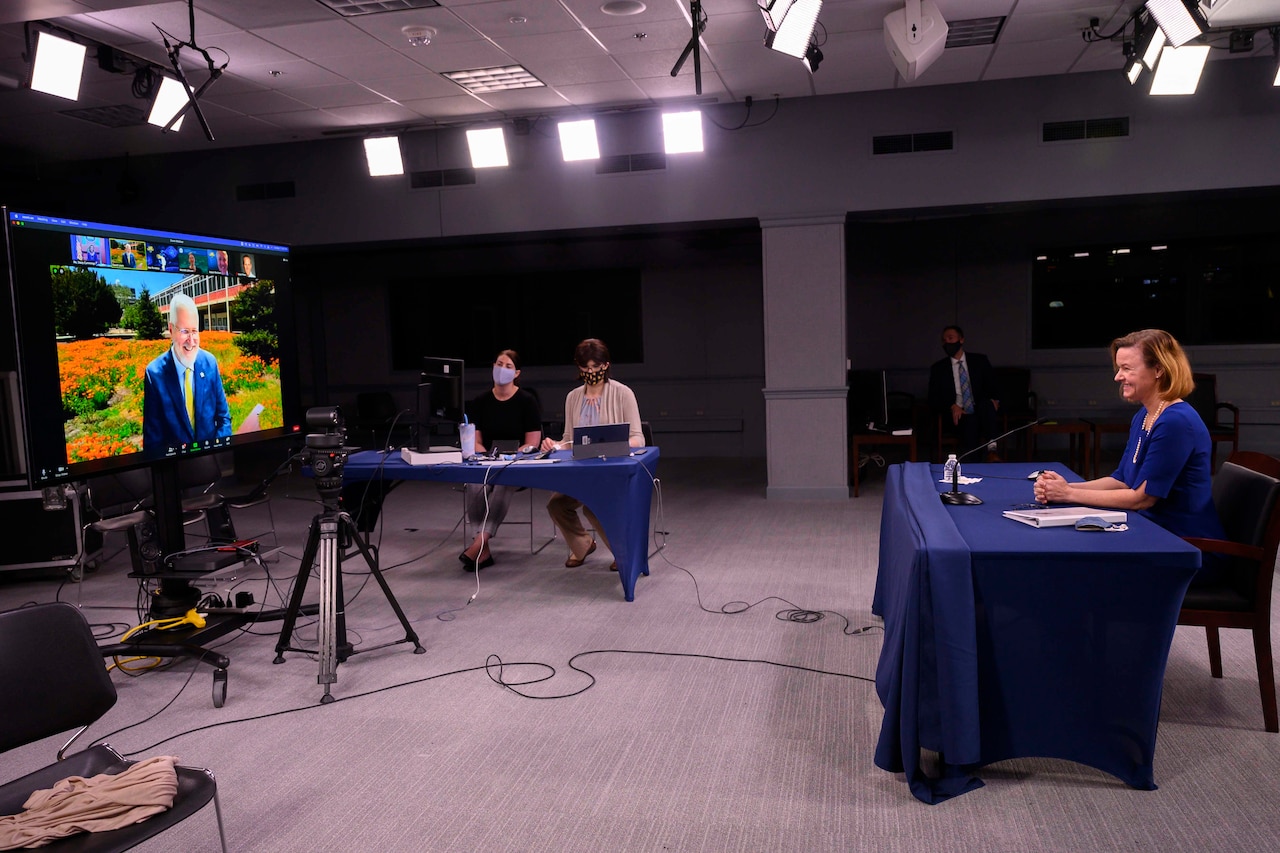 A woman sits at a table and looks at a large television screen. A video camera sits in front of the television screen and is pointed at the woman.