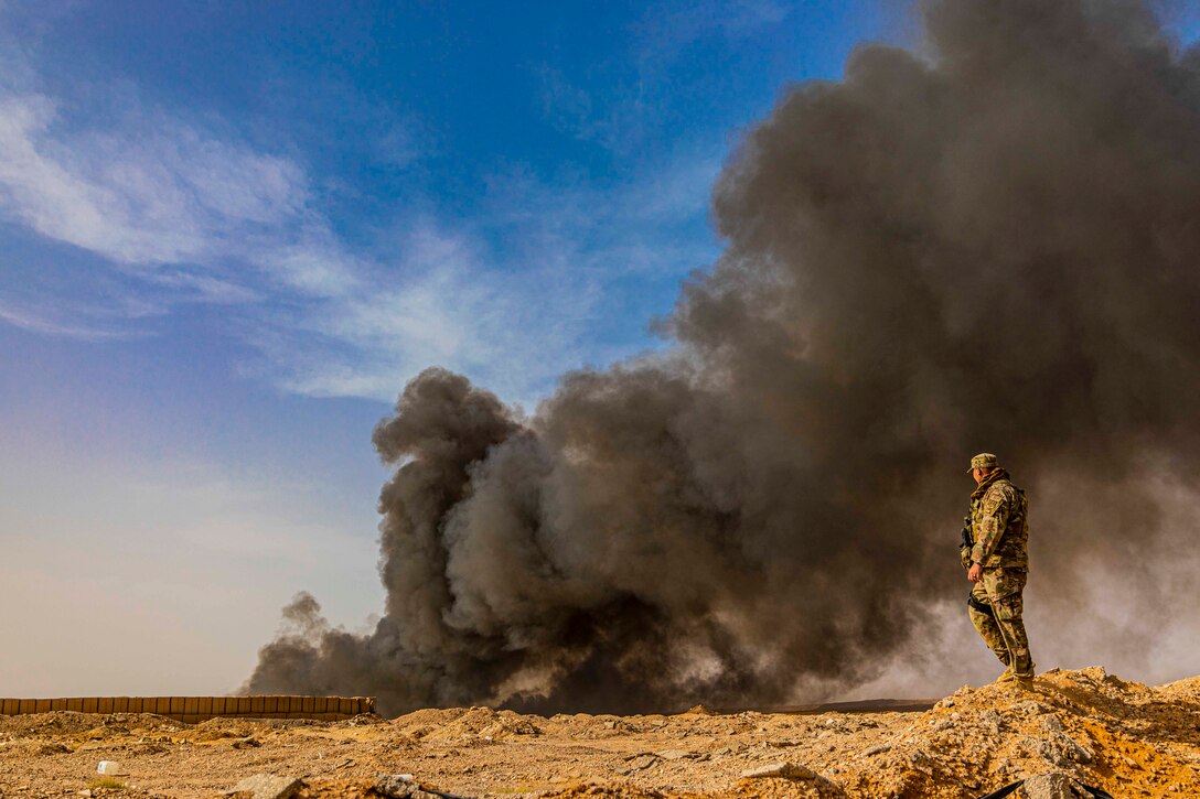 An airman stands on a dirt mound as black smoke rises in the distance.
