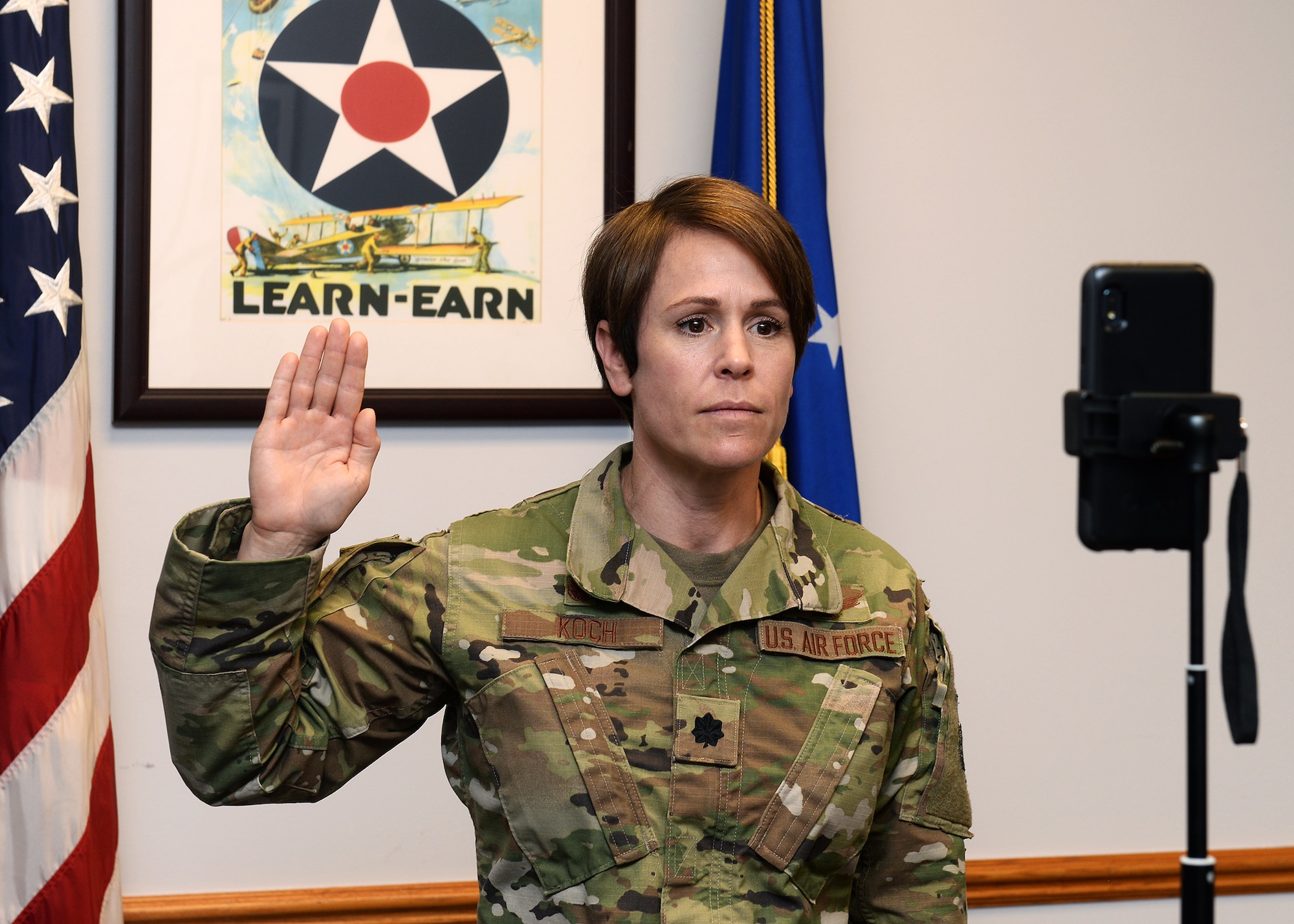 close up photograph of female lieutenant colonel in uniform with U.S. and Air Force flags behind her. Her right hand is raised as she stands in front of cell phone and verbally administers the oath of enlistment