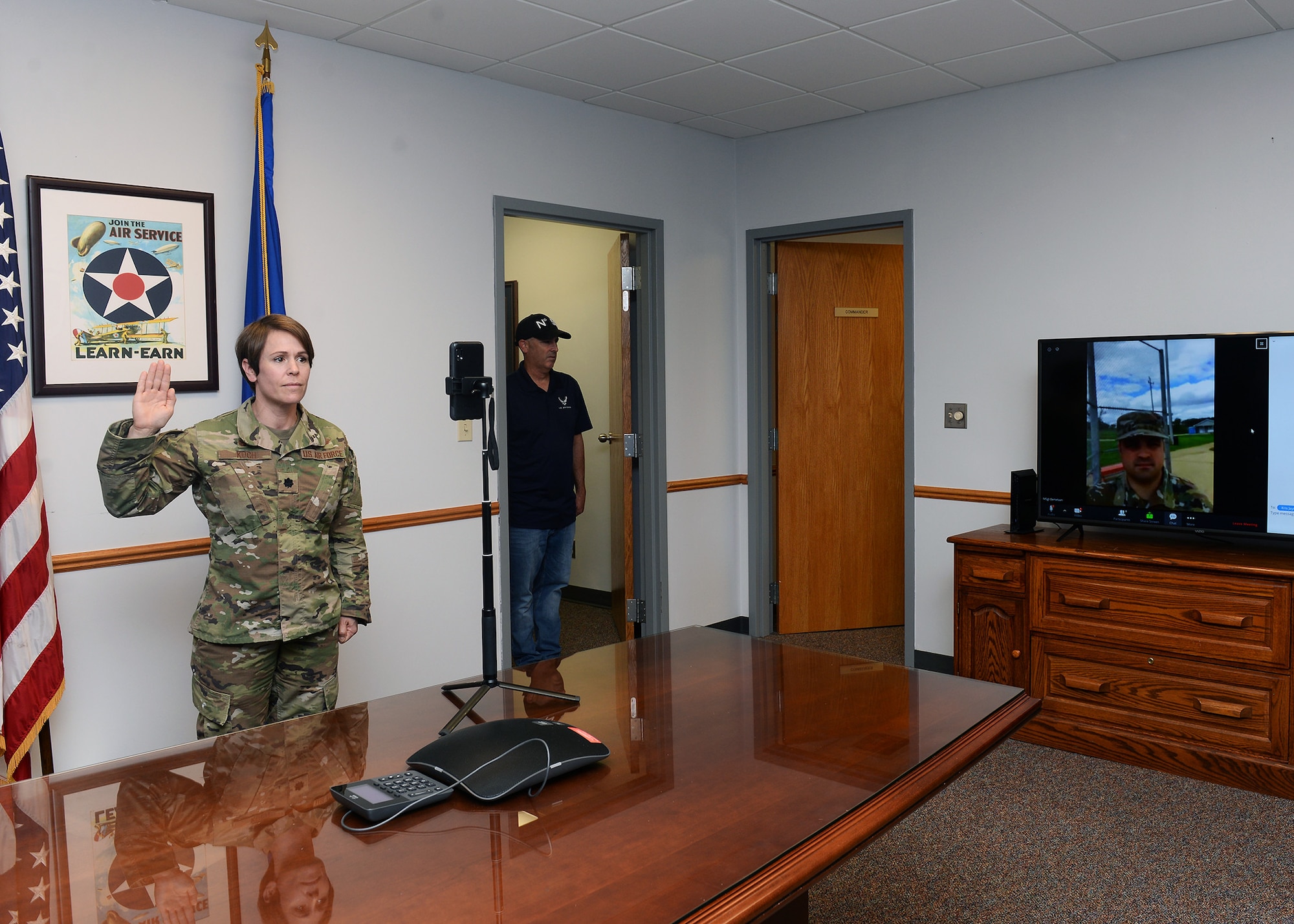 female Lieutenant Colonel standing in conference room with U.S. flag and Air Force flags behind her and in front of cell phone with her right hand raised to the right of the photograph a Technical Sergeant on a large television screen; as he takes oath of enlistment