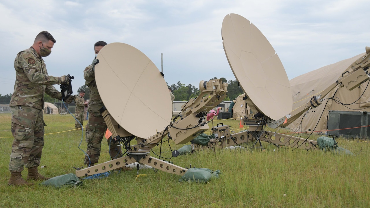 Airmen work on satellite dishes in a grassy field.