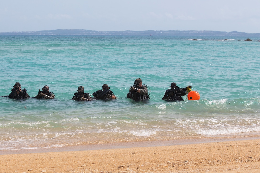 A line of six Marines move through water, one holding an orange item.