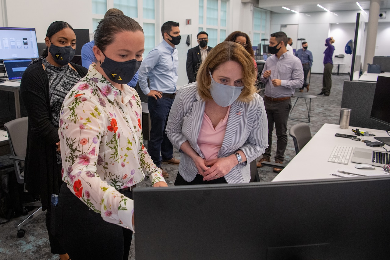 Two women look at a computer monitor in an office as others watch from behind.