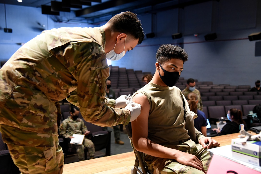 A guardsman wearing a face mask and gloves leans over to apply a bandage after giving a vaccine to a service member who is seated in a chair.