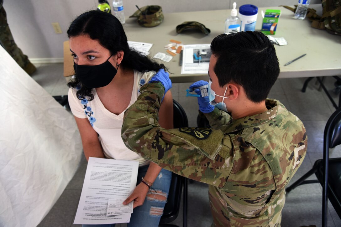 A soldier wearing a face mask and gloves gives a vaccine to a woman seated in a chair.