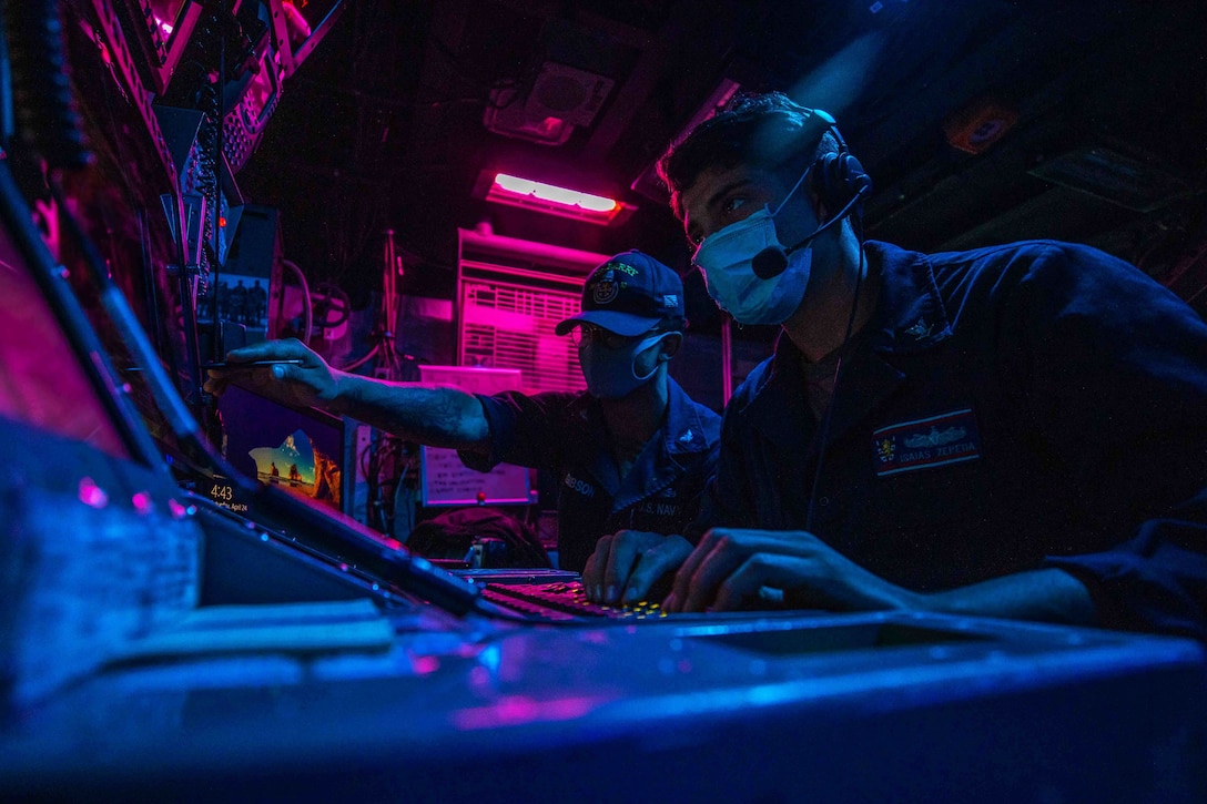 Two sailors monitor a board on a ship lit in blue and pink lights.