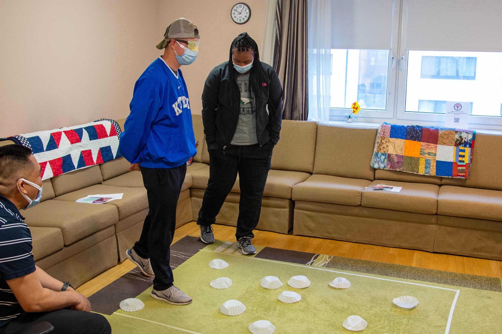 A woman guides a blindfolded man across a carpet with coffee filters on it.