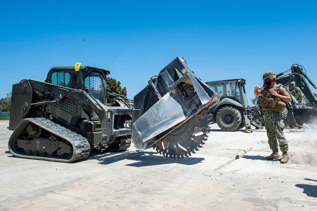 A sailor directs another operating a small construction vehicle.