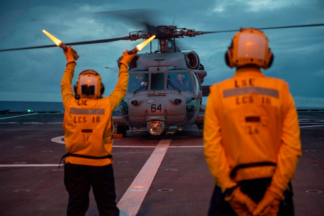 A sailor signals to a helicopter on a ship while a fellow sailor watches.