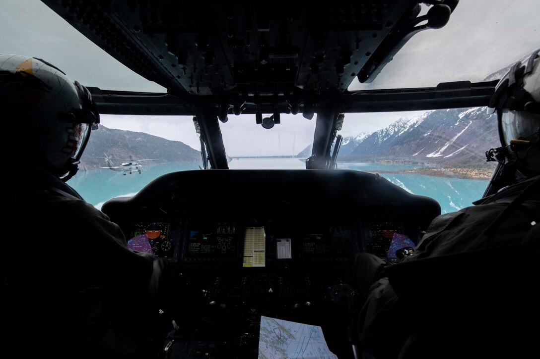 Two pilots sit in the cabin of a helicopter as they fly over water.