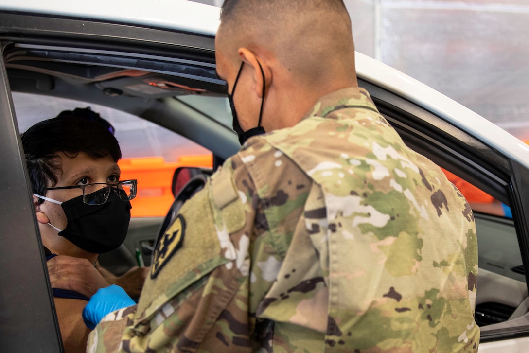 A soldier wearing a face mask and gloves administers a  vaccine to a resident while sitting in their car.
