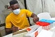 A servicemember sits in a yellow shirt in front of a table of red and blue testing vials. Covi