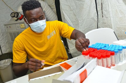 A servicemember sits in a yellow shirt in front of a table of red and blue testing vials. Covi