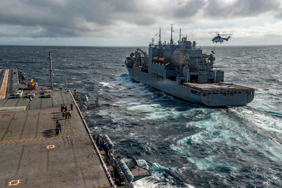 A helicopter carrying cargo on a sling-load hovers over one ship as another travels beside it.