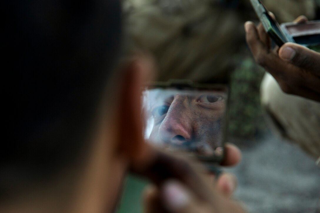 A Marine's painted face is visible in a mirror he's holding while applying camouflage paint, as other Marine holds a mirror nearby.