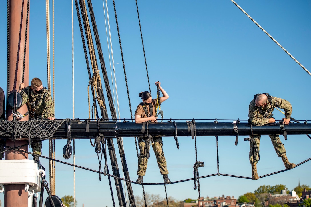 Three sailors stand on a line extending out from a ship's mast.