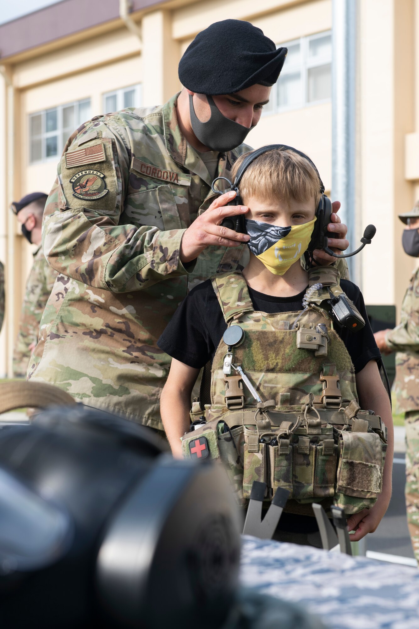 Tech. Sgt. Joshua Cordova, 374th Security Forces Squadron NCO in charge of training, helps Ayden Holt put on a pair of headphones at the “Day in the Life” event during National Police Week at Yokota Air Base, Japan, May 11, 2021.  During the event, the 374th SFS training section provided information on the non-lethal and breaching equipment they use. (U.S. Air Force photo by Staff Sgt. Joshua Edwards)