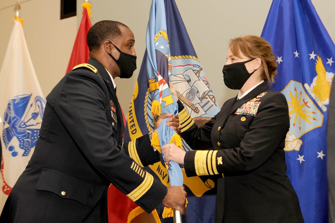 A man stands to left passing the DLA Troop Support organizational flag to a woman on the right. Both are in military service dress uniforms.
