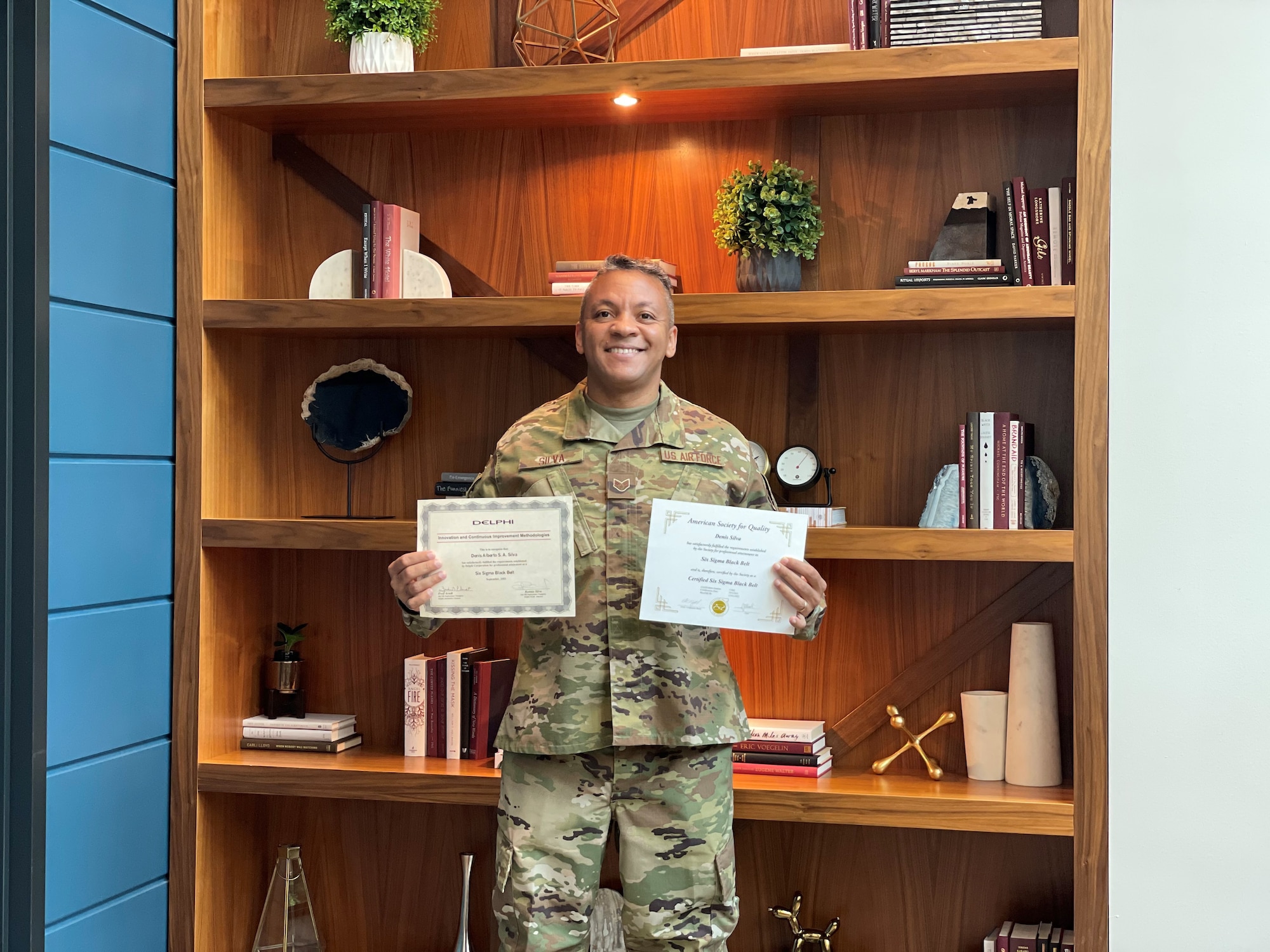 Staff Sgt. Denis Silva holds two certificates in front of a set of shelves.