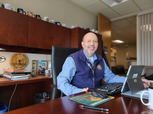 A man in a blue button up shirt and blue fleece vest sits at his desk in his office.