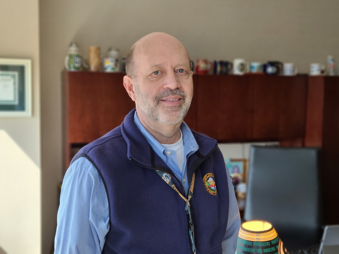 A man in a blue button up shirt and blue fleece vest stands in his office.