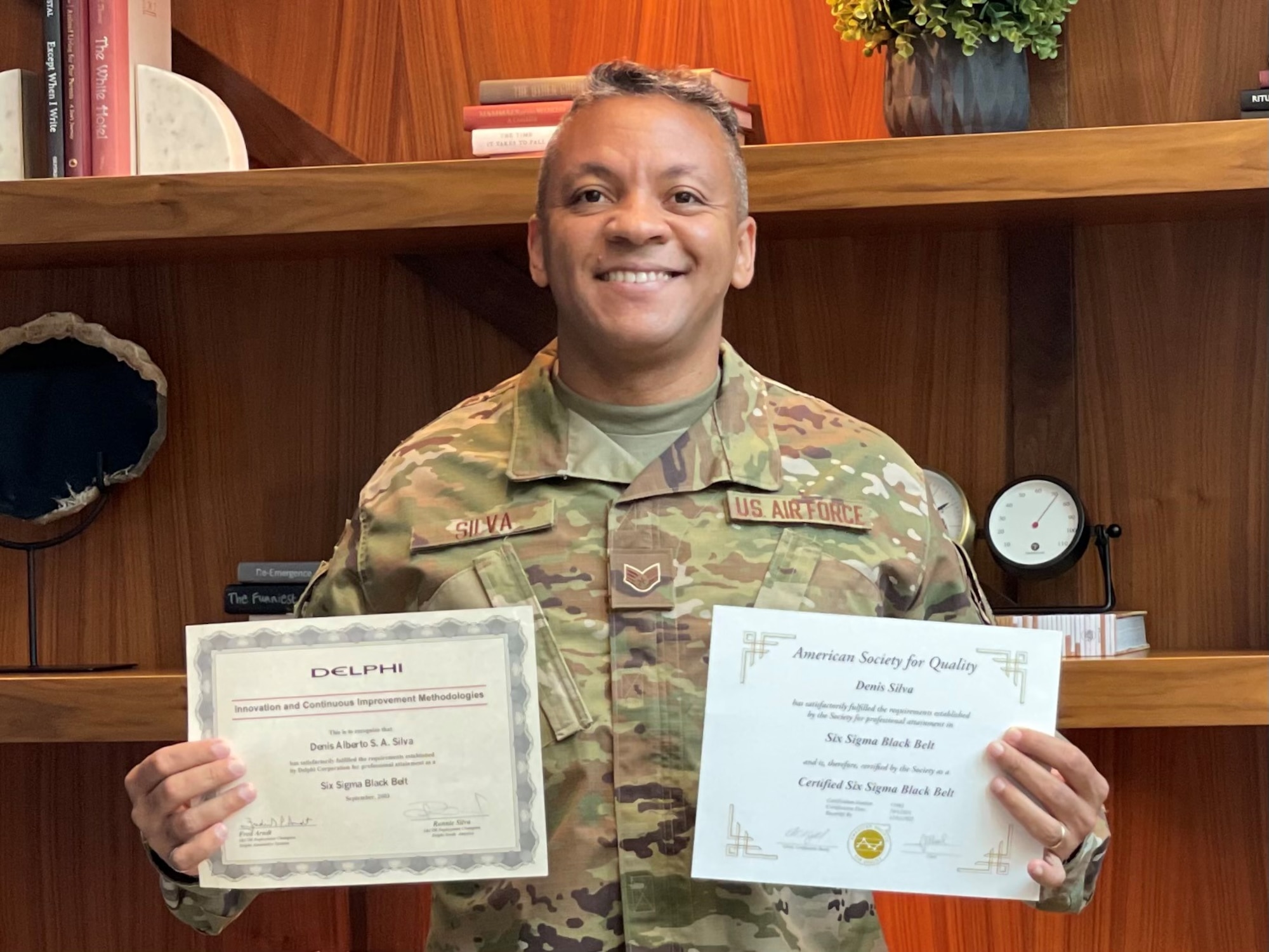 Staff Sgt. Denis Silva holds two certificates in front of a set of shelves.